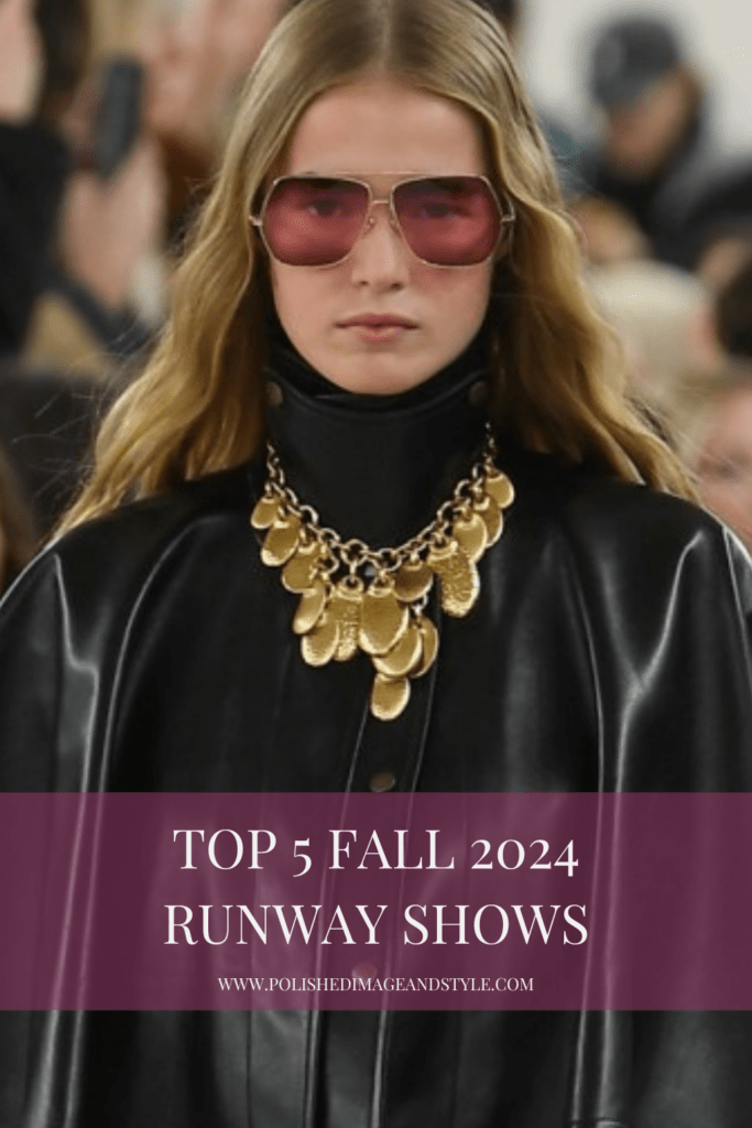 A woman with blonde hair wearing rose-colored glasses, a leather cape, and a gold necklace walks down the runway.