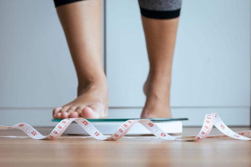 A woman steps on a scale with a measuring tape in front of her on the floor. 