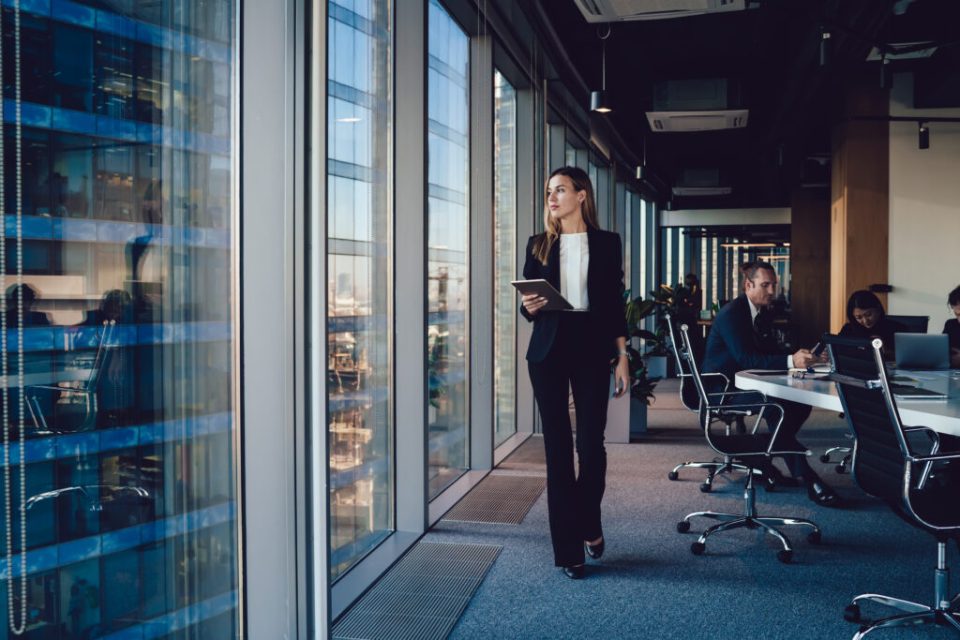 A woman walks along the side of an office looking out the window.