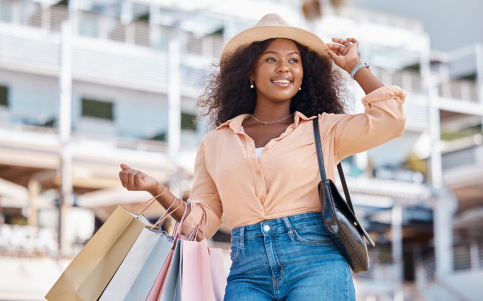 A woman with long curly hair holds her hat while wearing shopping bags on her arm. She is wearing a peach colored shirt.