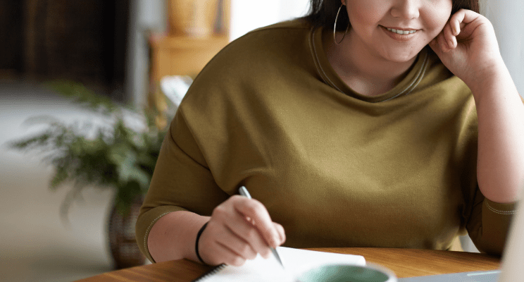 Woman in brown sweater smiling at table and writing with pen and notebook