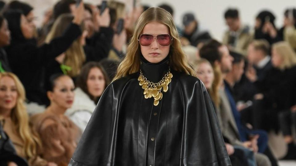 A woman with blonde hair wearing rose-colored glasses, a leather cape, and a gold necklace walks down the runway.